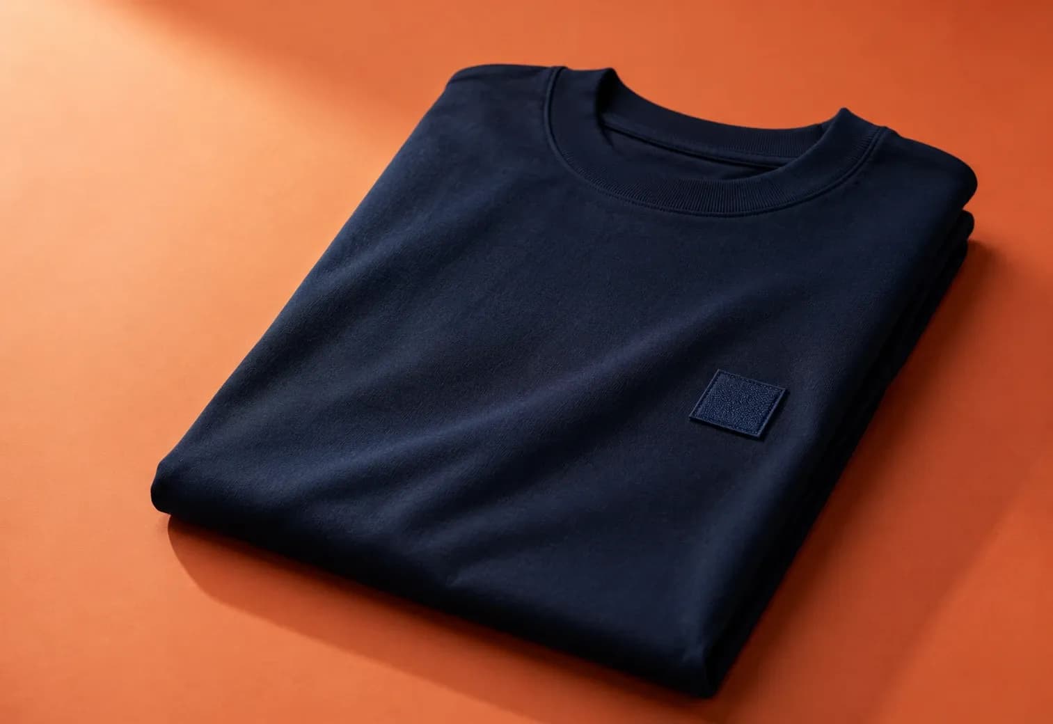 A folded navy t-shirt with a small square patch on the chest, lying on an orange surface lit from the upper left